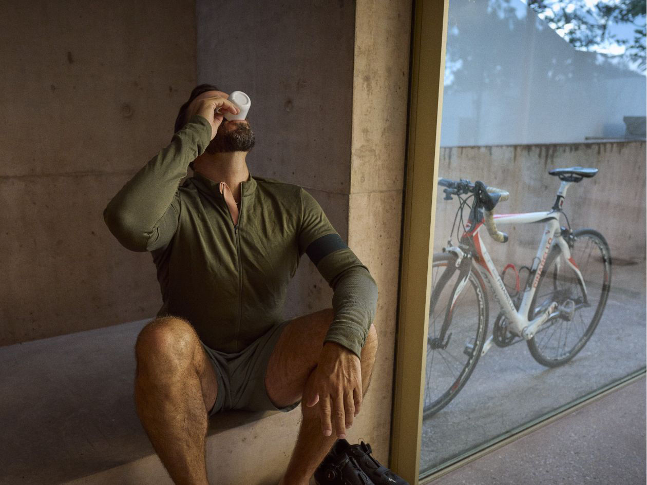 Man sitting indoors drinks from a cup beside a window with a bicycle outside